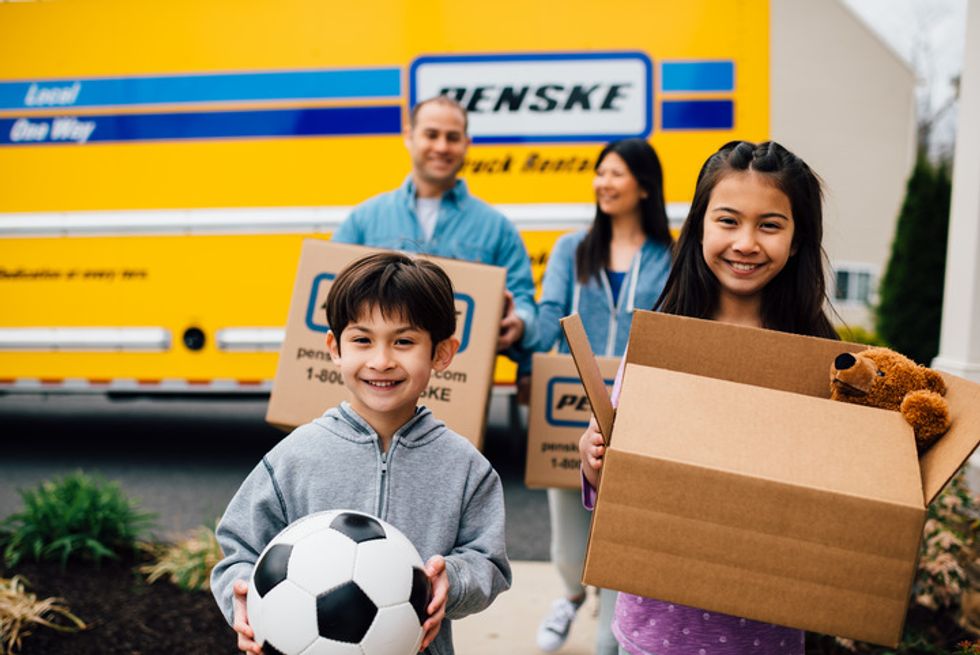 A family carries boxes and toys out of a yellow Penske moving truck.