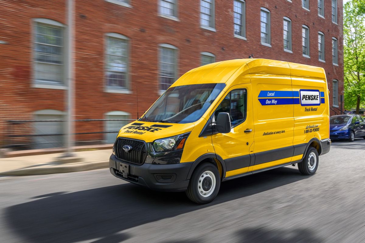 Yellow cargo van with Penske branding driving down street passing a brick building